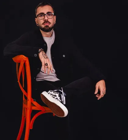 Luiz Dias sentado numa cadeira laranja, de jaqueta preta, fundo preto.